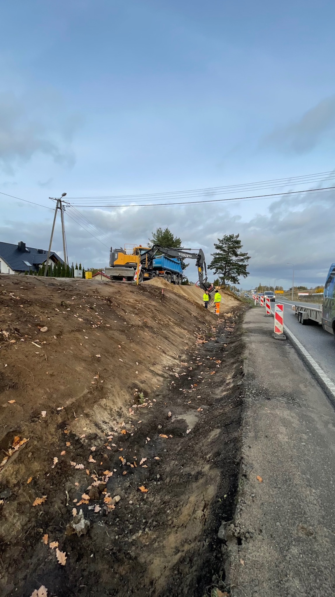Żółta koparka Volvo i wywrotka na placu budowy, dwóch pracowników w odblaskowych kamizelkach, widok na drogę i domy w tle, pochmurne niebo.