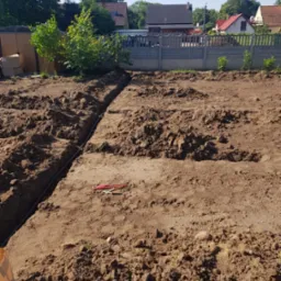 Wykopane rowy w ziemi pod instalację w ogrodzie, widoczne narzędzia, w tle domy i ogrodzenie.