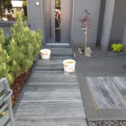 Nowoczesne wejście do domu z szarą elewacją, wyłożone płytami chodnikowymi z jasnymi fugami, obok rosną iglaki i małe drzewko, widoczne dwa wiadra z zawartością, prawdopodobnie do fugowania.