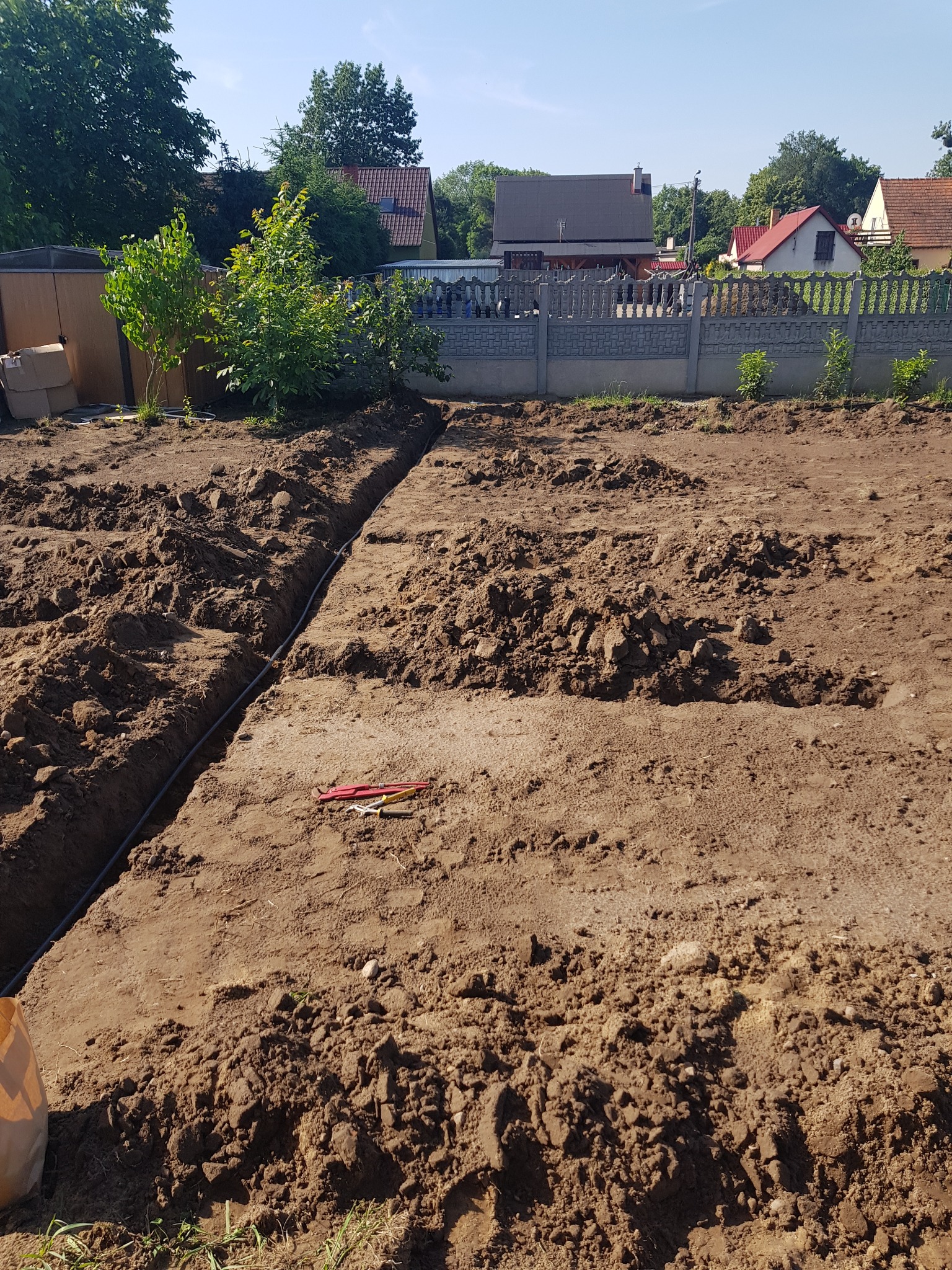 Wykopane rowy w ziemi pod instalację w ogrodzie, widoczne narzędzia, w tle domy i ogrodzenie.