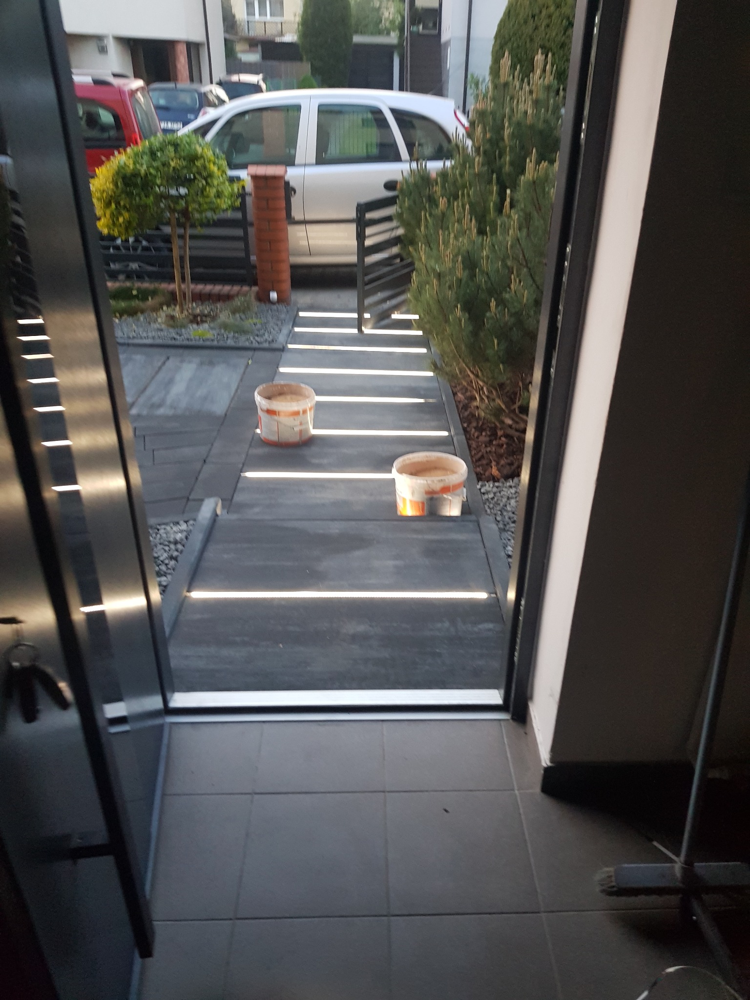 Nowoczesne podejście do domu z podświetlanymi płytami chodnikowymi, widoczne dwa wiadra farby i elementy zieleni w tle.