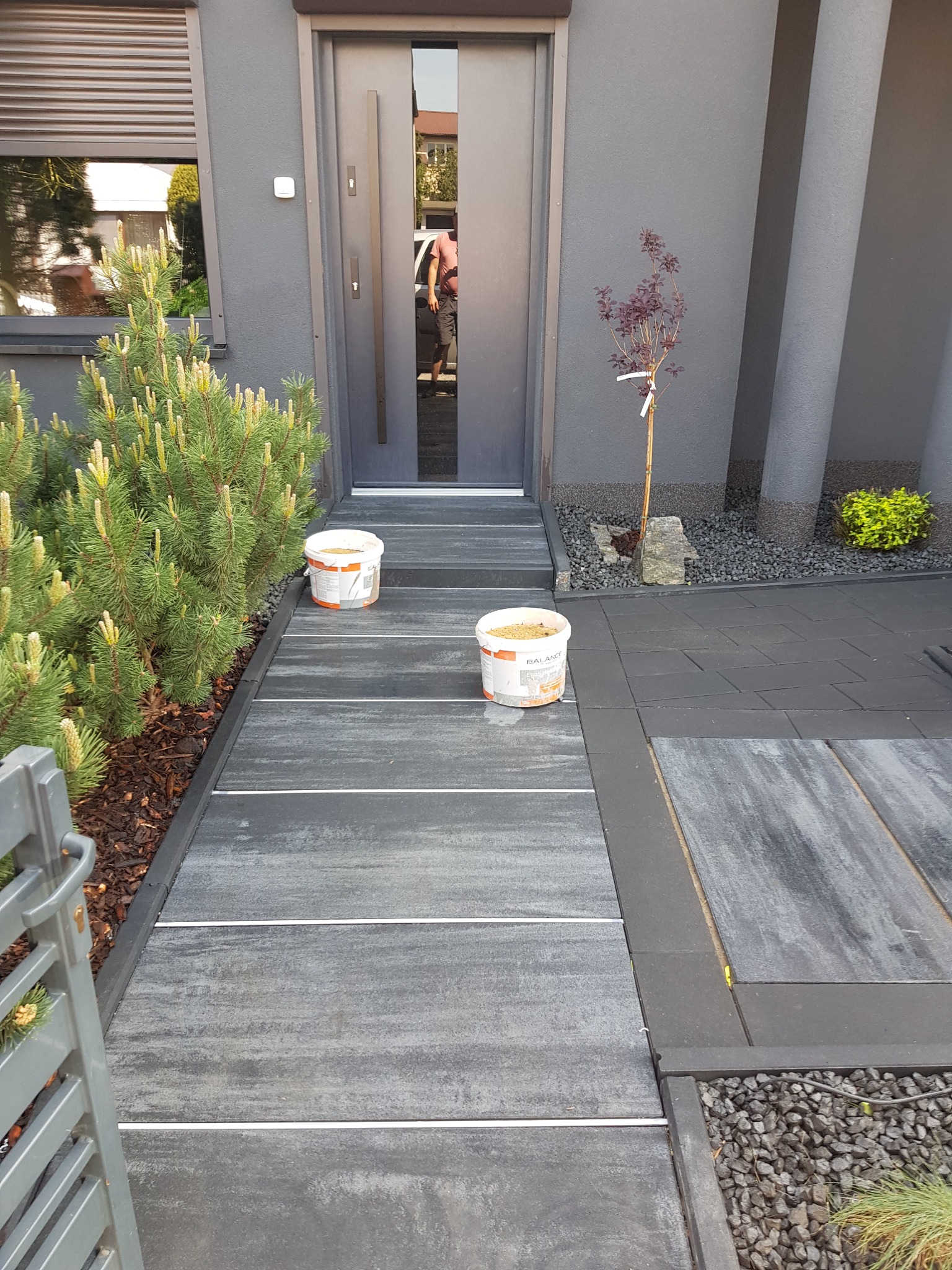Nowoczesne wejście do domu z szarą elewacją, wyłożone płytami chodnikowymi z jasnymi fugami, obok rosną iglaki i małe drzewko, widoczne dwa wiadra z zawartością, prawdopodobnie do fugowania.