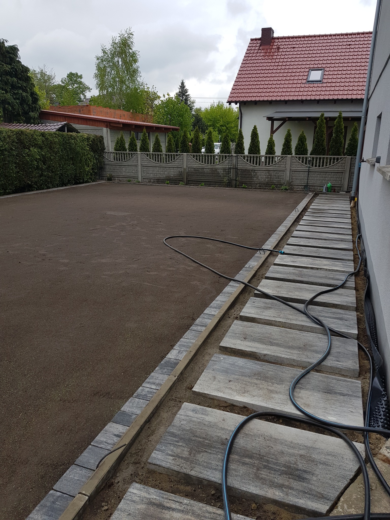 Przygotowany teren pod trawnik z rolki, obrzeża z kostki, ścieżka z płyt kamiennych i zwinięty wąż ogrodowy na posesji z domem w tle.