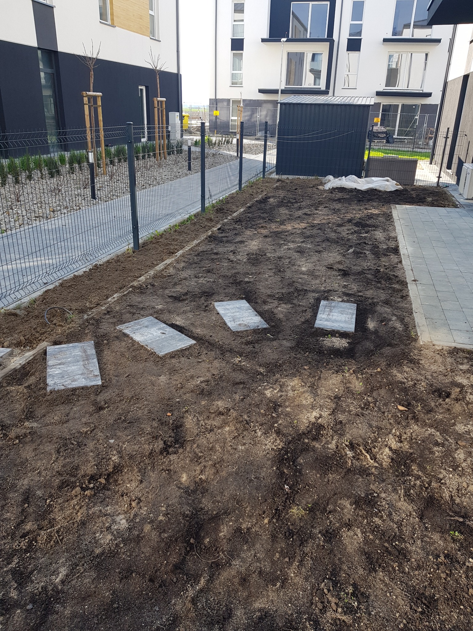 Przygotowany teren pod ogród z ułożonymi płytami chodnikowymi, widoczne budynki mieszkalne w tle i siatka ogrodzeniowa.
