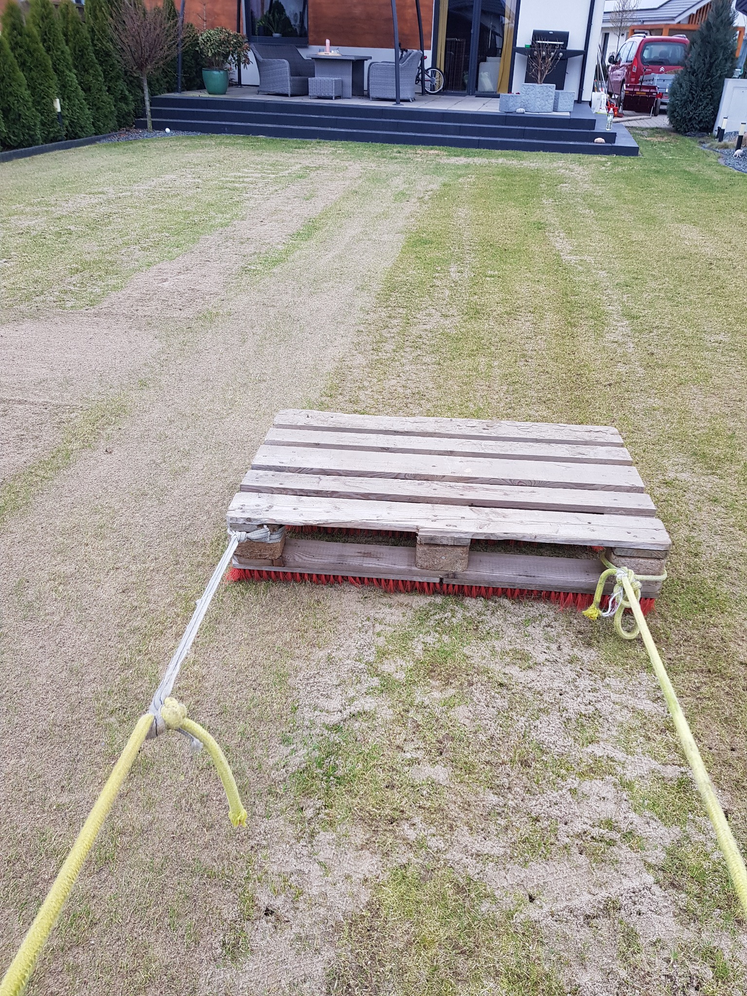 Paleta z przymocowaną szczotką ciągnięta po trawniku pokrytym piaskiem; widoczne ślady po przejeździe, w tle dom z tarasem i krzewami.