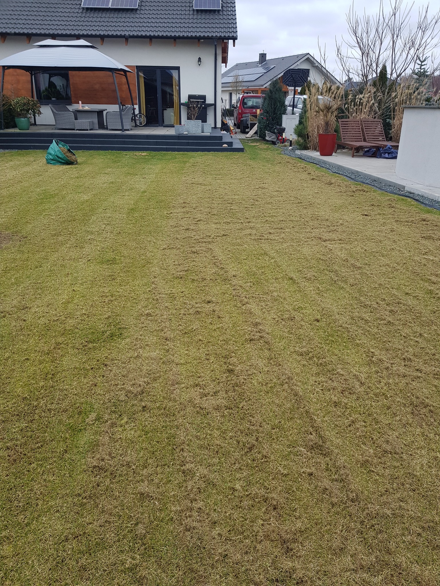 Rozległy trawnik po wertykulacji, z widocznymi resztkami organicznymi, w tle nowoczesny dom z panelami słonecznymi i elementami wyposażenia ogrodu.