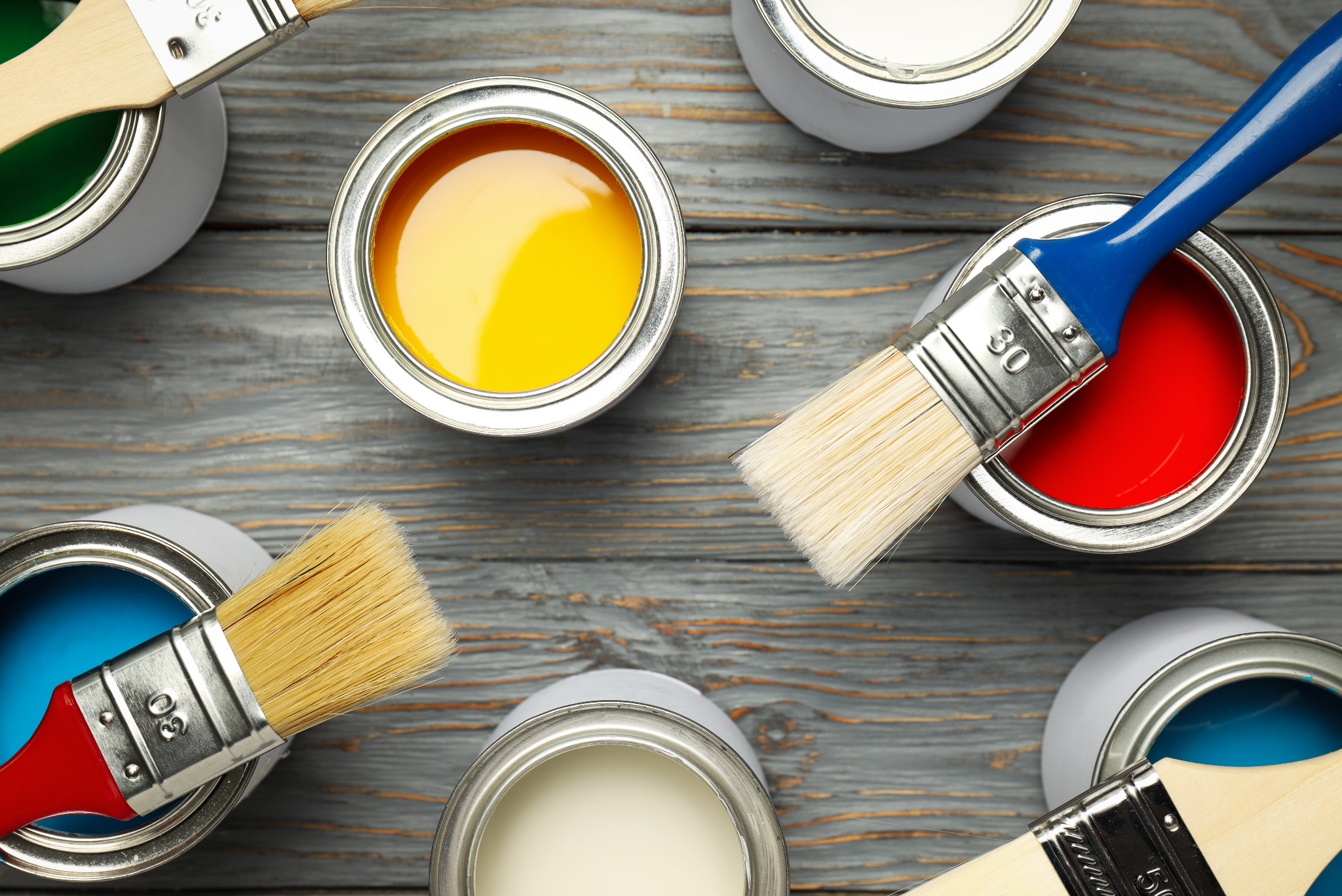 Zestaw otwartych puszek z farbami w kolorach: zielonym, żółtym, czerwonym, niebieskim i białym, każda z pędzlem, ułożonych na szarym, drewnianym tle.