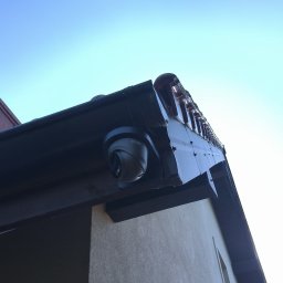 INSTA-GUARD'S - Kamera monitoringu marki DAHUA zamontowana pod okapem domu w Przyłękach, widok z dołu na tle błękitnego nieba.