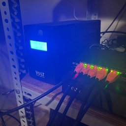 INSTA-GUARD'S - Zasilacz awaryjny UPS Volt Polska z wyświetlaczem napięcia, podłączony do switcha sieciowego pomarańczowymi kablami ethernet, widoczne diody sygnalizacyjne.