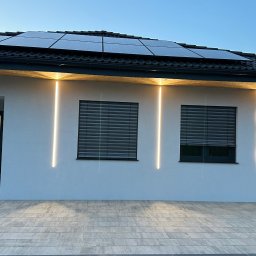 INSTA-GUARD'S - Nowoczesny dom z panelami słonecznymi na dachu i minimalistycznym oświetleniem elewacyjnym w postaci pionowych, wąskich pasków LED po bokach okien z roletami zewnętrznymi.