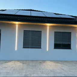 Nowoczesny dom z panelami słonecznymi na dachu i minimalistycznym oświetleniem elewacyjnym w postaci pionowych, wąskich pasków LED po bokach okien z roletami zewnętrznymi.