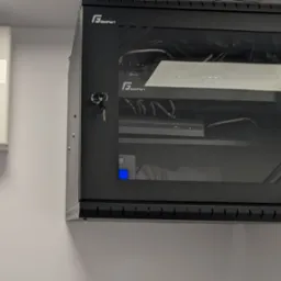 Zamknięta szafa rack GetFort z widocznymi urządzeniami sieciowymi (router TP-Link, rejestrator) i kablami, zamontowana na ścianie obok białej obudowy Satel Integra 64.