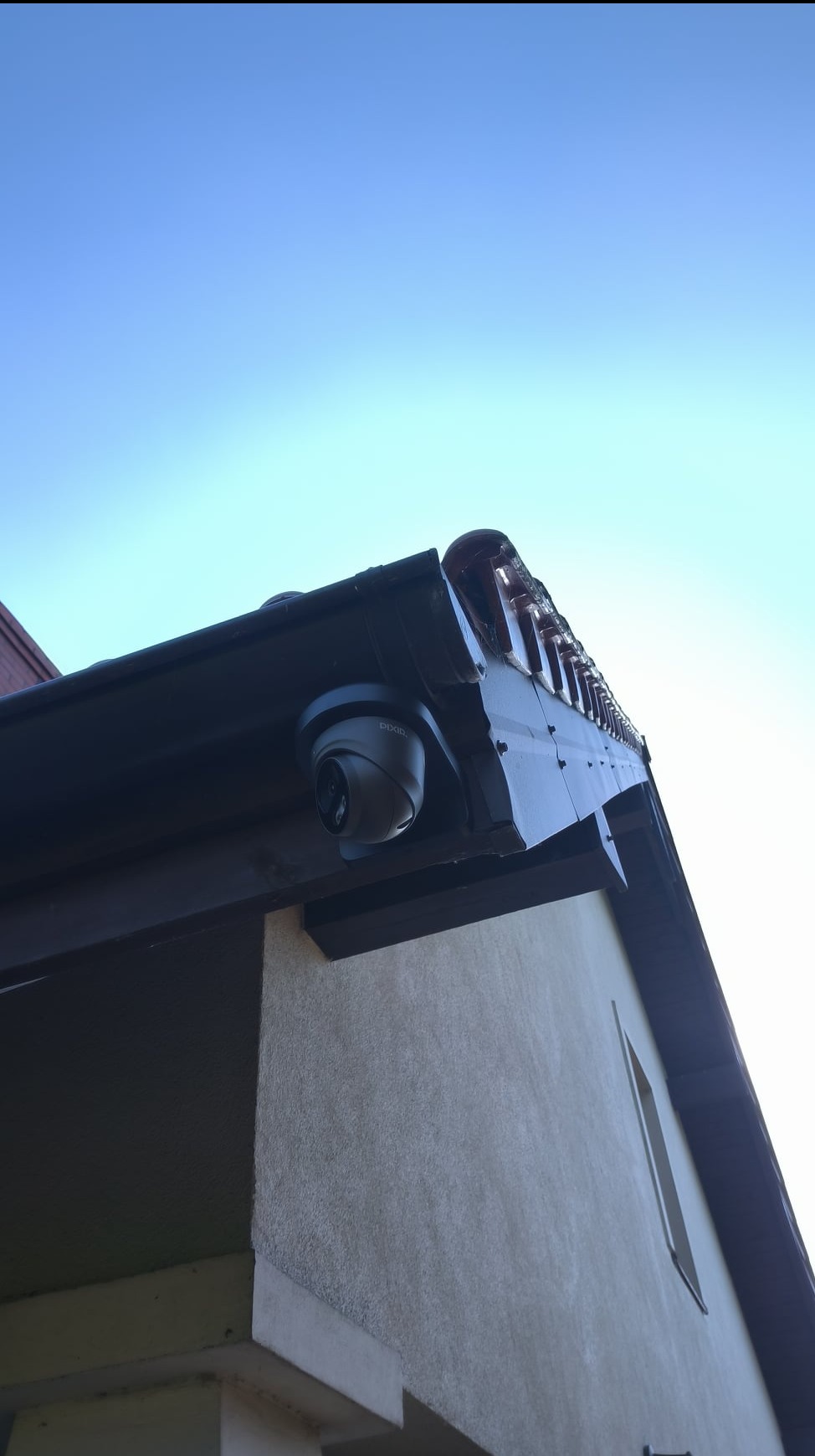 Kamera monitoringu marki DAHUA zamontowana pod okapem domu w Przyłękach, widok z dołu na tle błękitnego nieba.