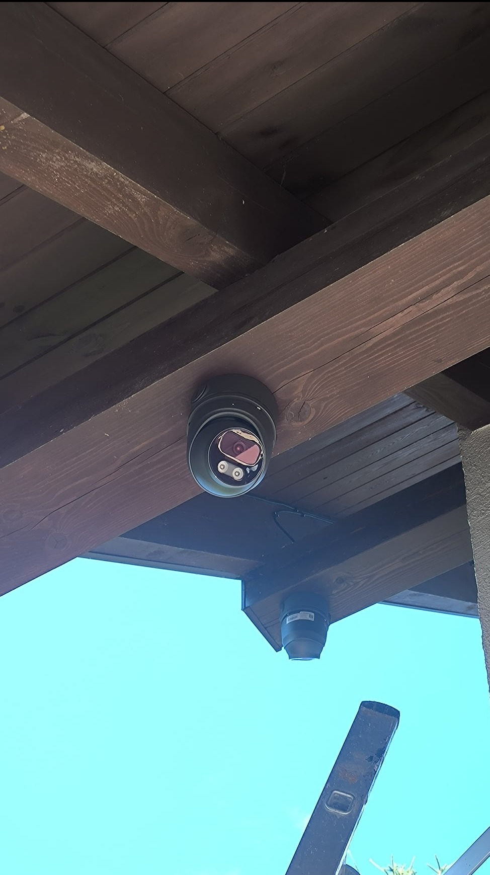 Kamera kopułkowa zamontowana na drewnianej konstrukcji dachu, widok z dołu na tle błękitnego nieba, fragment drabiny w kadrze.