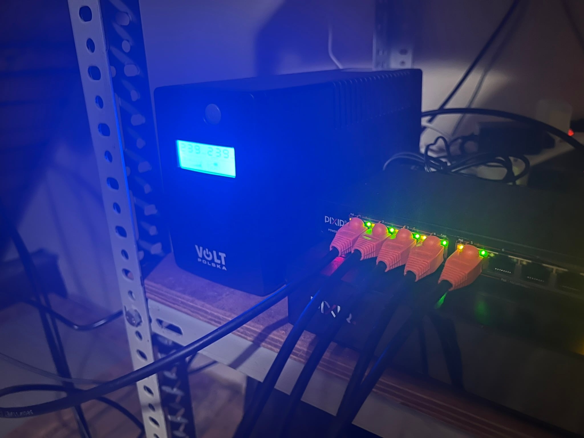 Zasilacz awaryjny UPS Volt Polska z wyświetlaczem napięcia, podłączony do switcha sieciowego pomarańczowymi kablami ethernet, widoczne diody sygnalizacyjne.