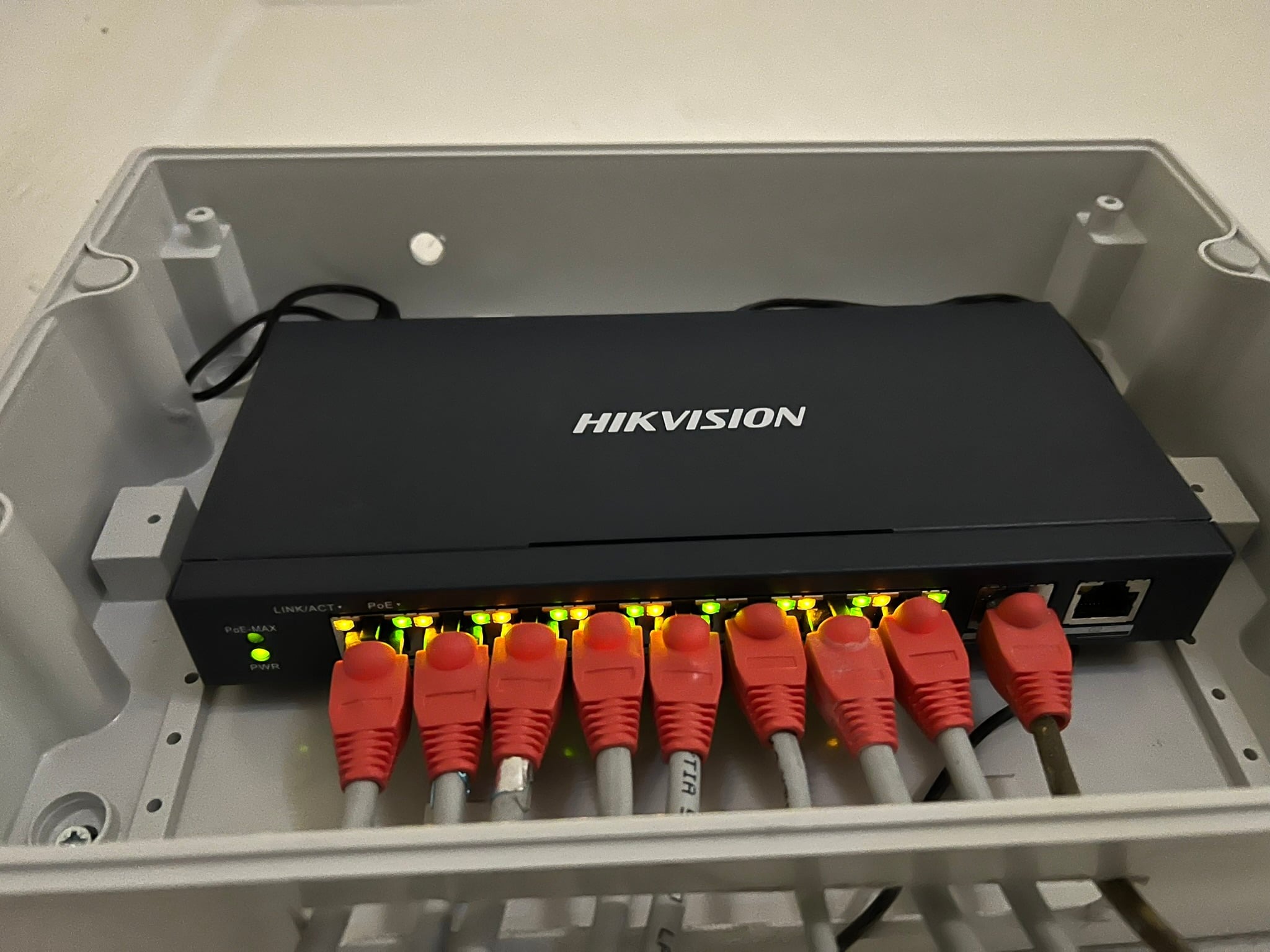 Switch Hikvision w szarej obudowie z podłączonymi czerwonymi kablami Ethernet i świecącymi diodami LED.