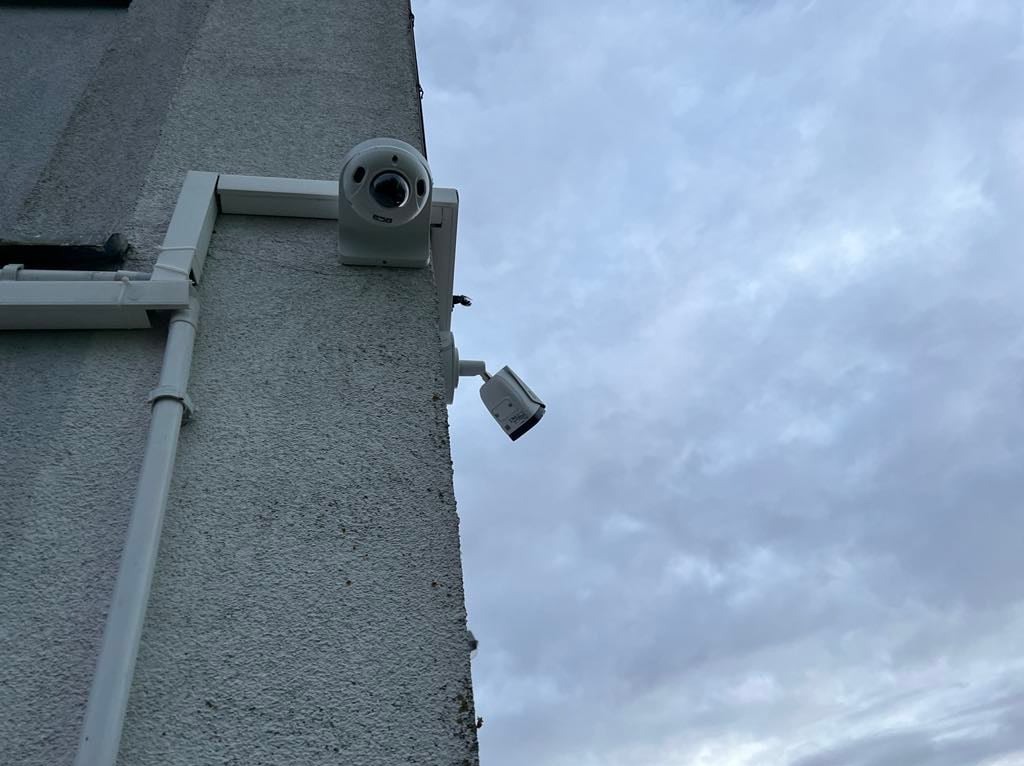 Dwie kamery monitoringu zamontowane na rogu budynku z szorstką, jasną elewacją, widoczne rury spustowe i pochmurne niebo w tle.