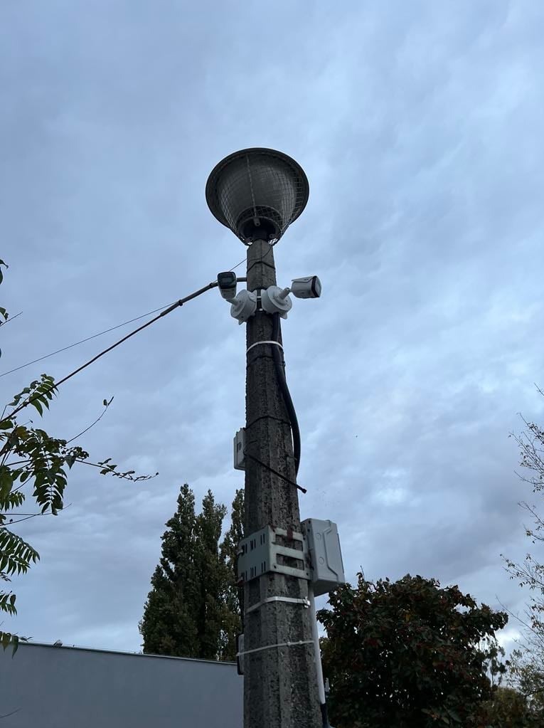 Betonowy słup oświetleniowy z zamontowanymi kamerami CCTV i puszkami instalacyjnymi, widok z dołu na tle pochmurnego nieba.