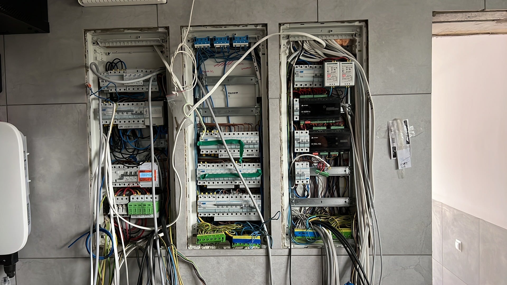 Trzy otwarte skrzynki elektryczne w ścianie, widoczne liczne przewody, bezpieczniki i moduły, instalacja w trakcie podłączania, na ścianie notatka z numerem.