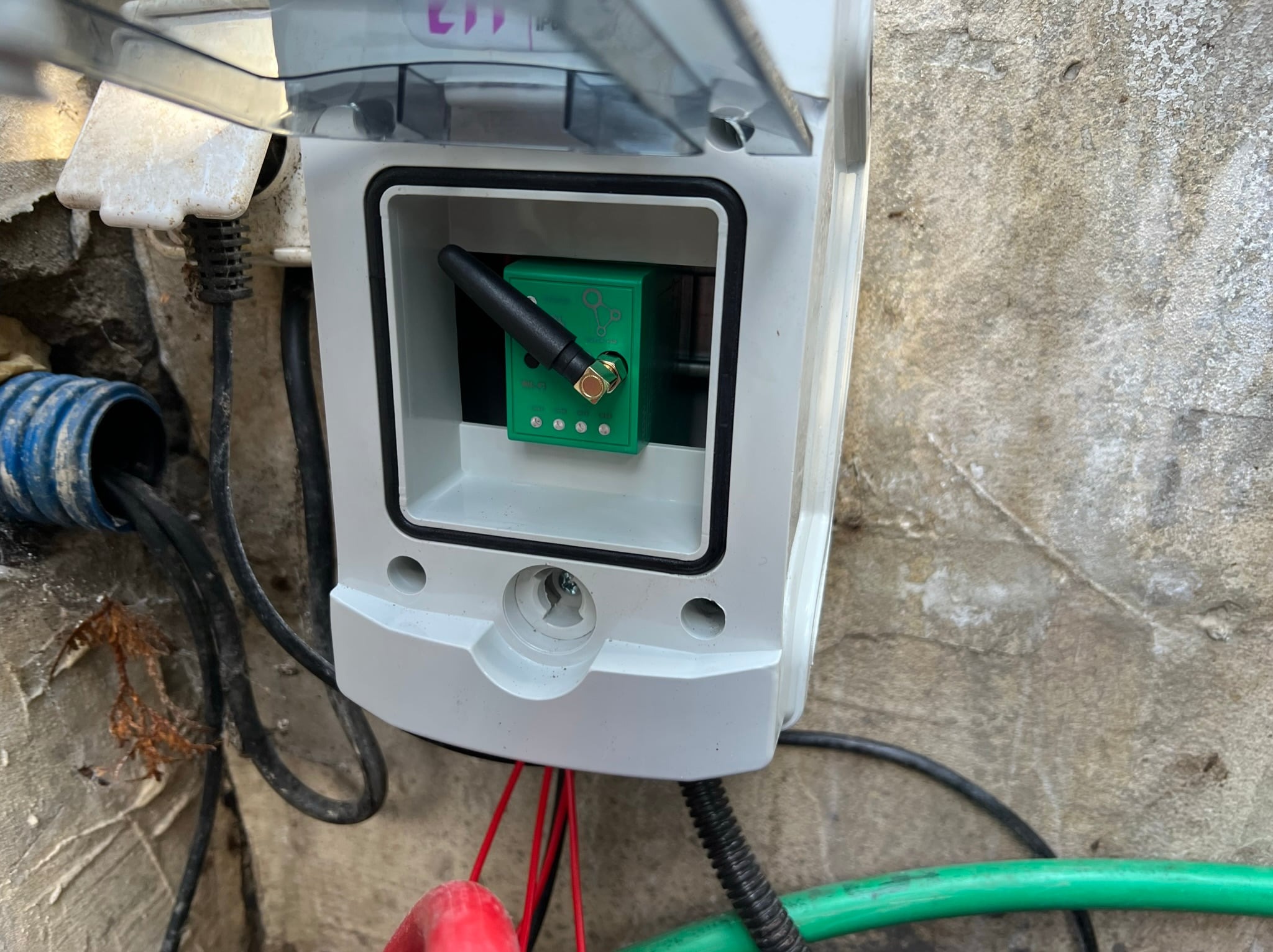 Otwarta skrzynka elektryczna z zielonym modułem sterującym i anteną na szarej ścianie, widoczne kable i rury.