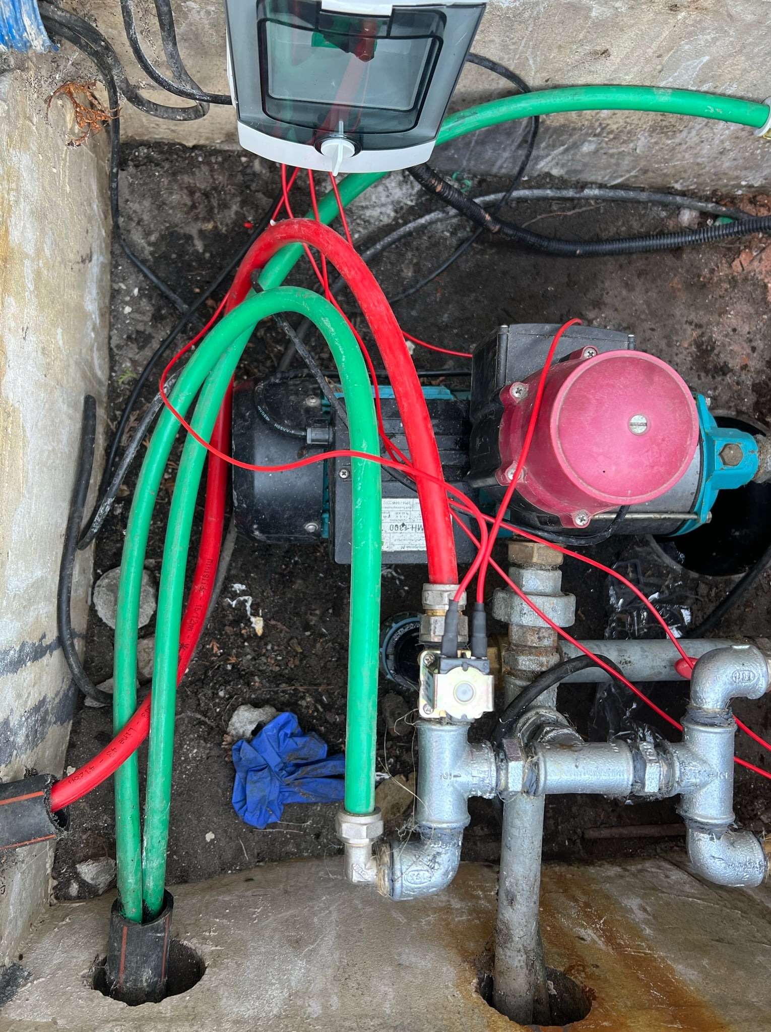Instalacja hydroforu w studzience: widoczne rury doprowadzające wodę, pompa hydroforowa z czerwonym zbiornikiem ciśnieniowym, zawory, przewody elektryczne oraz skrzynka elektryczna. W tle widoczne...