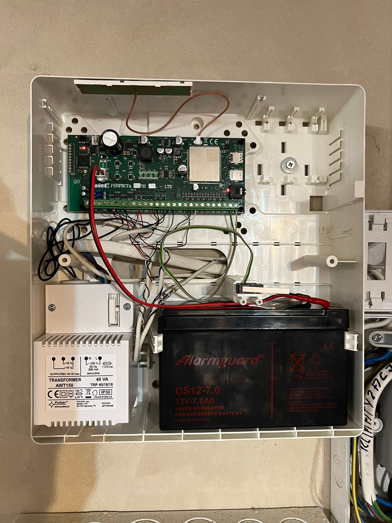Otwarta obudowa centrali alarmowej z widoczną elektroniką, akumulatorem Alarmguard, transformatorem Pulsar AWT150 i plątaniną przewodów. Widoczne elementy montażowe i fragment ściany w tle.