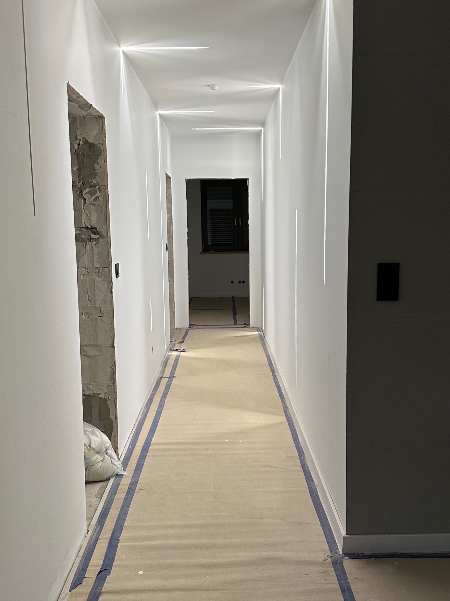 Długi, wąski korytarz w trakcie wykańczania; widoczne białe ściany z liniowym oświetleniem LED, zabezpieczona podłoga, surowy otwór drzwiowy z workiem gruzu oraz fragment ciemnej ściany.