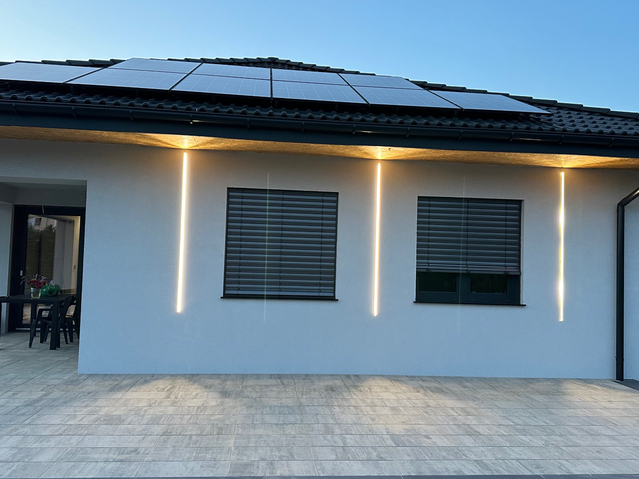 Nowoczesny dom z panelami słonecznymi na dachu i minimalistycznym oświetleniem elewacyjnym w postaci pionowych, wąskich pasków LED po bokach okien z roletami zewnętrznymi.