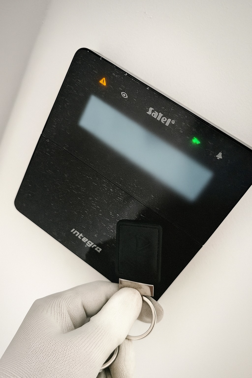 Panel alarmowy Satel Integra z diodami LED, obsługiwany przez dłoń w białej rękawiczce za pomocą breloka zbliżeniowego. Widoczne logo Satel.