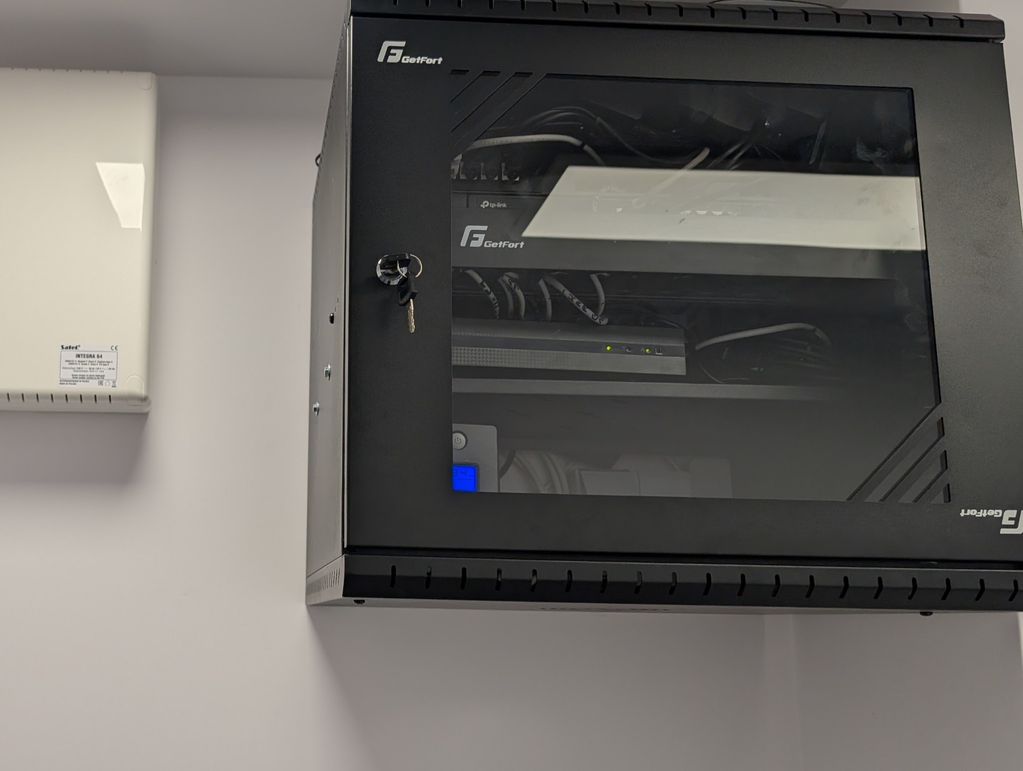 Zamknięta szafa rack GetFort z widocznymi urządzeniami sieciowymi (router TP-Link, rejestrator) i kablami, zamontowana na ścianie obok białej obudowy Satel Integra 64.