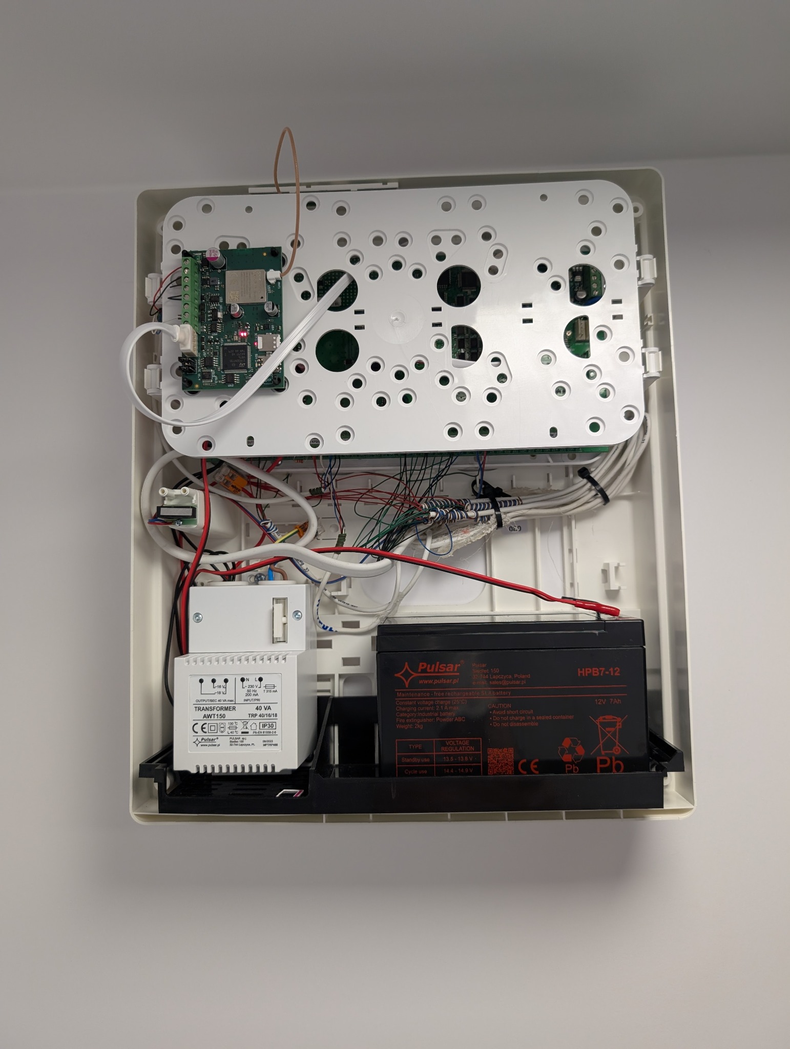 Otwarta obudowa centrali alarmowej z widocznymi elementami elektroniki, transformatorem i akumulatorem. Instalacja wykonana wewnątrz białej obudowy na ścianie.