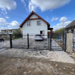 ROMBUD - Nowo zamontowana, czarna, metalowa brama wjazdowa z ogrodzeniem panelowym przed domem jednorodzinnym z kostką brukową, widoczny samochód za bramą i robotnik przy krawężniku po lewej stronie.