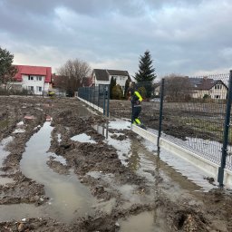 ROMBUD - Montaż ogrodzenia panelowego na grząskim terenie po deszczu; widoczny pracownik w odblaskowej kamizelce i betonowa podmurówka.