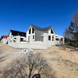 ROMBUD - Wznoszenie ogrodzenia z szarej bloczków betonowych wokół nowoczesnego domu z szarą dachówką, na placu budowy pod błękitnym niebem.