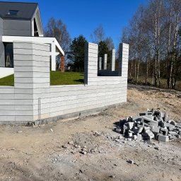 ROMBUD - Wznoszona konstrukcja z szarych, betonowych bloczków ogrodzeniowych, z widocznym stosem elementów do budowy, na tle nowoczesnego domu i drzew bezlistnych.