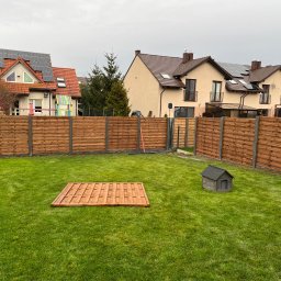 ROMBUD - Nowoczesne, poziome drewniane ogrodzenie panelowe oddzielające zadbany trawnik z domkiem dla psa i pojedynczym panelem ogrodzeniowym leżącym na trawie od zabudowy jednorodzinnej.