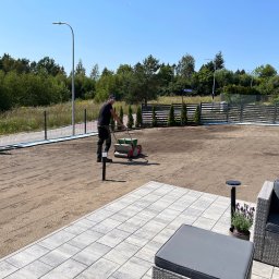 ROMBUD - Pracownik w czarnym stroju rozprowadza nasiona trawy siewnikiem na przygotowanym podłożu, widoczne świeżo posiane ślady, w tle nowy metalowy płot i młode tuje, z przodu szara kostka brukowa tarasu...