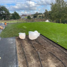 ROMBUD - Świeżo ułożona trawa z rolki w Gdańsku, widoczne elementy systemu nawadniania w trakcie instalacji oraz przygotowane do montażu lampy ogrodowe w foliowych osłonach.