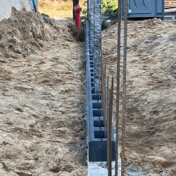 ROMBUD - Wykop z ułożonymi bloczkami betonowymi tworzącymi fundament pod ogrodzenie, z wystającymi prętami zbrojeniowymi, na tle ziemi i hydrantu.