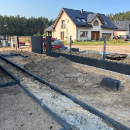 ROMBUD - Wykop pod fundament ogrodzenia z betonowych paneli, widoczne przygotowane słupki, w tle dom jednorodzinny z panelami słonecznymi.