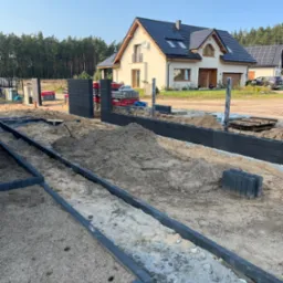 Wykop pod fundament ogrodzenia z betonowych paneli, widoczne przygotowane słupki, w tle dom jednorodzinny z panelami słonecznymi.