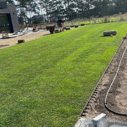 ROMBUD - Dwaj mężczyźni układają rolki trawy na przygotowanym podłożu, widoczny trawnik, wał do trawy, granica trawnika i nowoczesny dom w tle.