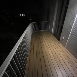 ROMBUD - Nocne ujęcie balkonu z drewnianą podłogą i białymi, metalowymi barierkami, widok na ciemny krajobraz z odległymi światłami.