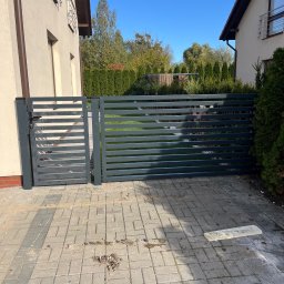 ROMBUD - Nowoczesna, grafitowa brama posesyjna z poziomych profili, widok z zewnątrz na kostkę brukową i zieleń ogrodu w tle.