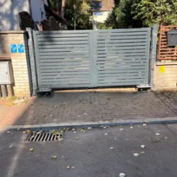 Nowoczesna, szara brama przesuwna z poziomych paneli, zamontowana na ceglanych słupkach, widok z zewnątrz na chodnik i ulicę z opadłymi liśćmi.