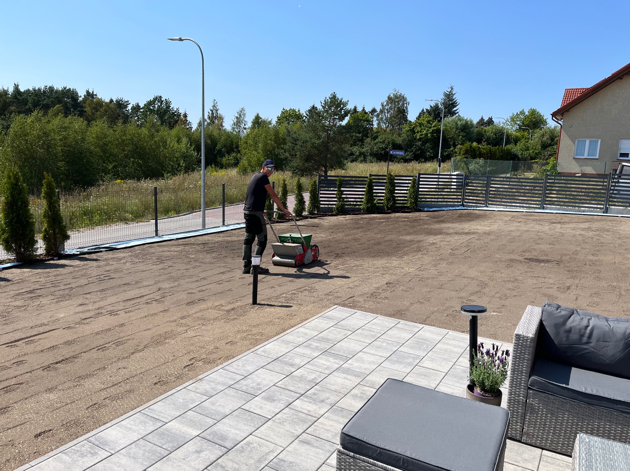 Pracownik w czarnym stroju rozprowadza nasiona trawy siewnikiem na przygotowanym podłożu, widoczne świeżo posiane ślady, w tle nowy metalowy płot i młode tuje, z przodu szara kostka brukowa tarasu...