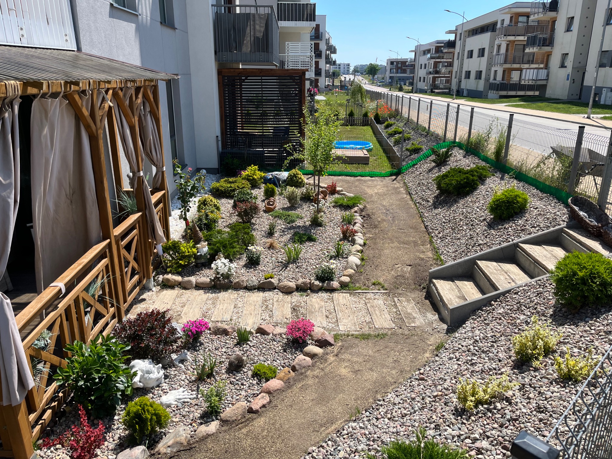 Widok z góry na zagospodarowany ogród przydomowy z drewnianą altaną z zasłonami, ścieżką z płyt chodnikowych, rabatami z kamyków, różnorodną roślinnością, małym basenem i ogrodzeniem siatkowym...