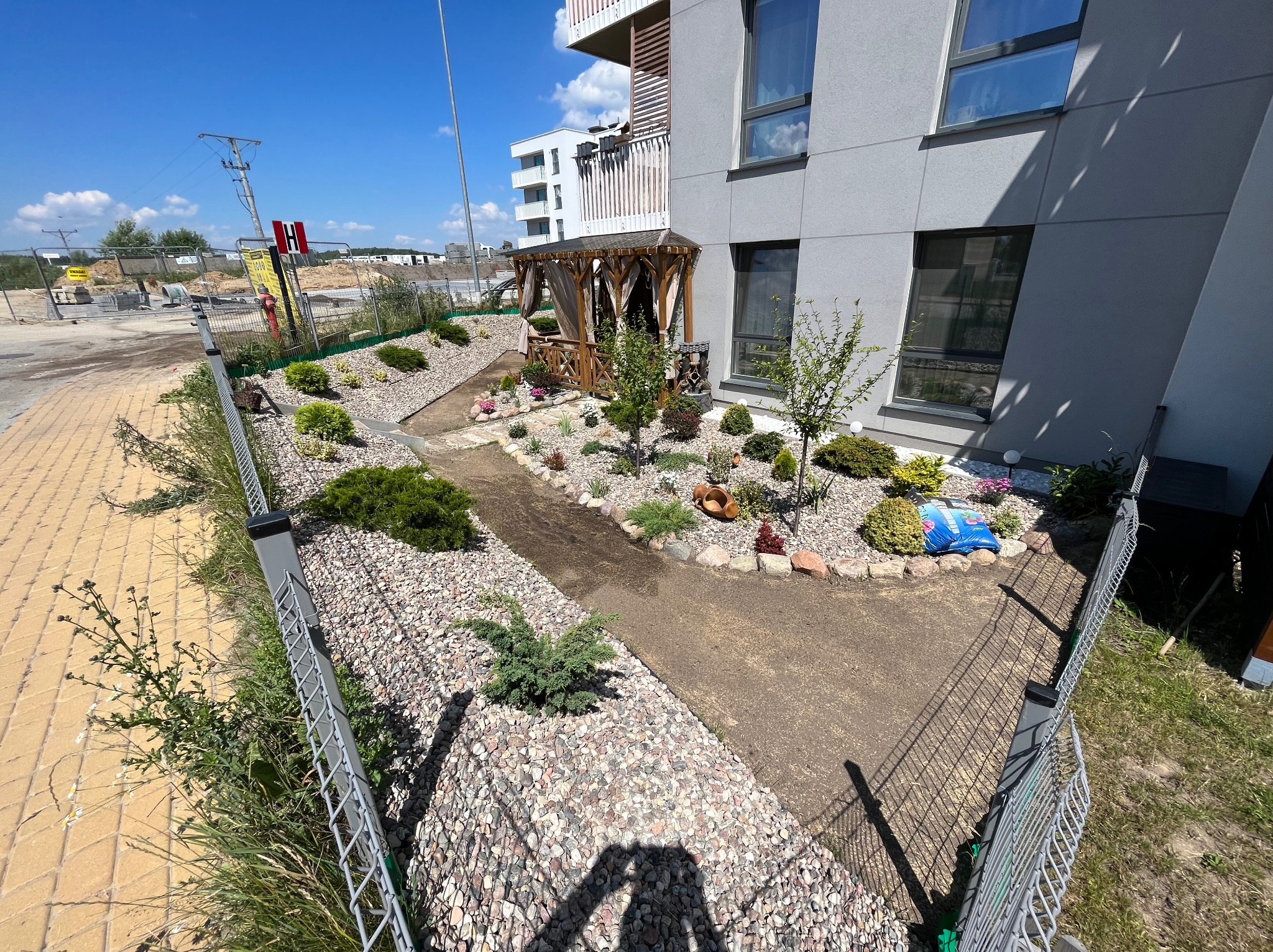 Nowoczesny ogród przy apartamentowcu z drewnianą altaną, ścieżką wysypaną kamieniami, ogrodzeniem z siatki i elementami małej architektury