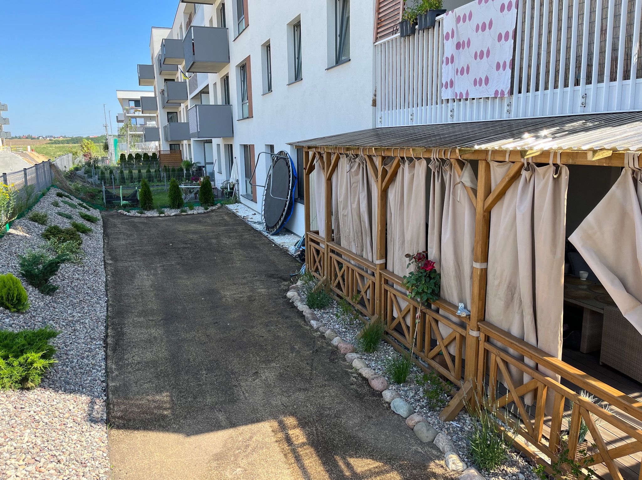 Teren przed budynkiem mieszkalnym po przygotowaniu pod trawnik z rolki, widoczne wyrównane podłoże, ozdobne krzewy i drewniana altana z zasłonami.