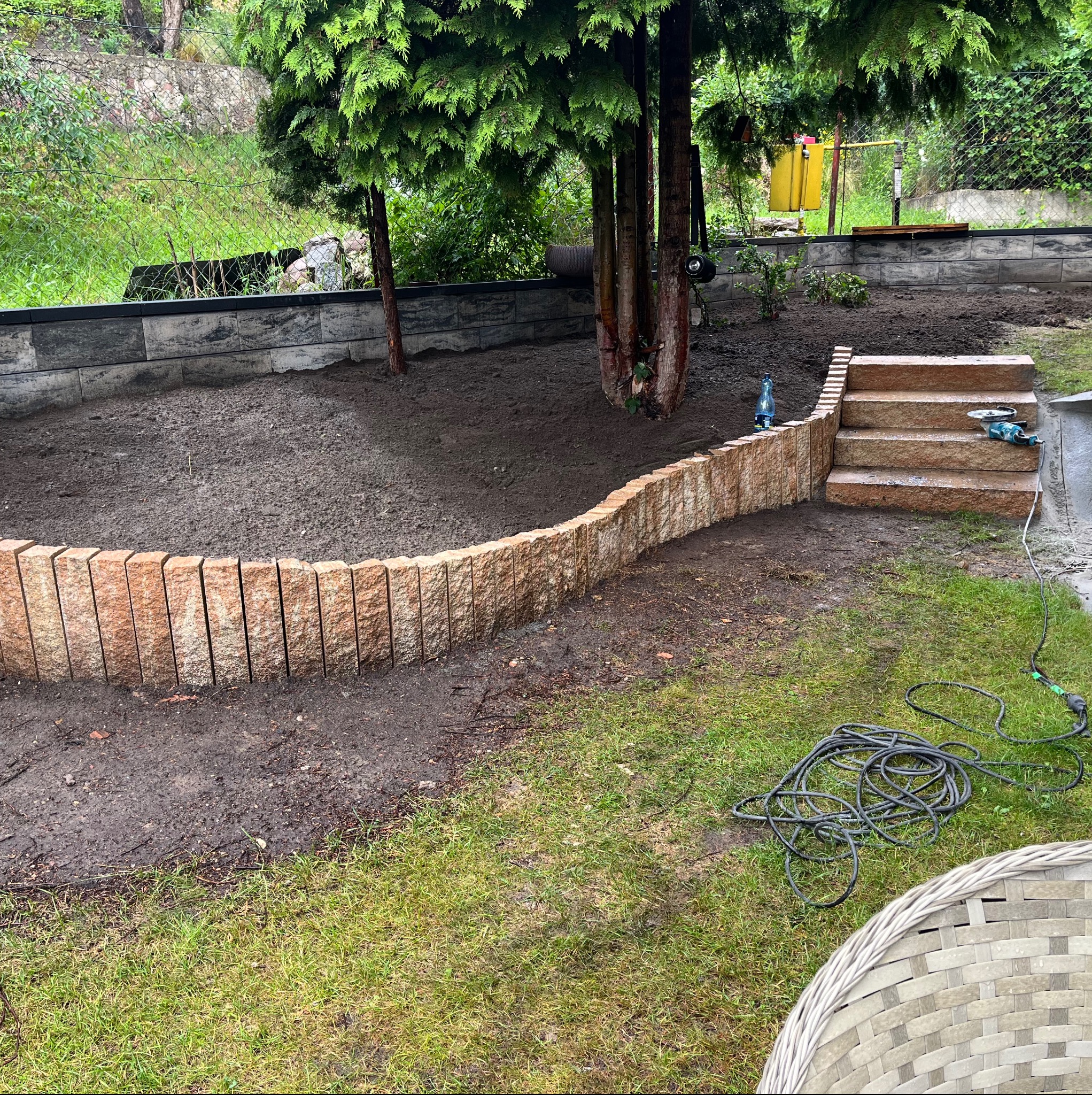 Kaskadowy ogród w trakcie budowy: obrzeże z pionowo ułożonych kamieni oddziela rabatę od trawnika, kamienne schody prowadzą na wyższy poziom, w tle drzewa iglaste.